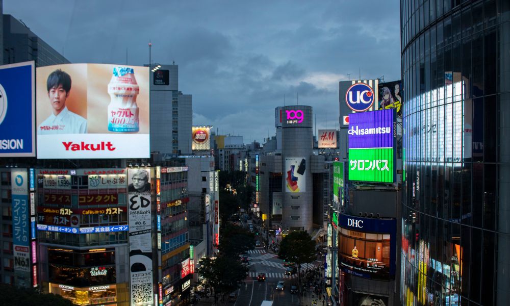 Se muestra la calle de una ciudad con cartelería exterior y pantallas de grandes dimensiones, además de mostrar cantantes con sus discos y próximas giras